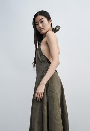 Noor naine, kellel on pikk tume juuksed, kannab õlavaba oliivrohelist kleiti punutud detailide ja avatava seljaga, seisab tavalise tausta ees.