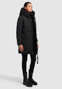 Svart pufferjacka med huva, med ett chevronmönster, dragkedjeslutning, sidofickor och en märkesetikett. Kombinerad med svarta leggings och stövlar.