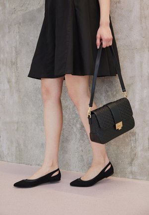 Bolso negro con un patrón texturizado y herrajes dorados, combinado con zapatillas de ante negras que presentan detalles recortados y punteras puntiagudas.
