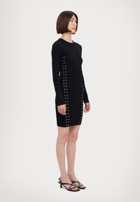 Black ribbed knit dress with long sleeves, featuring gold eyelets along the sides. It has a fitted shape and a minimalist design.