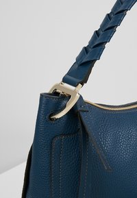 Sac à main en cuir texturé bleu foncé avec bandoulière tressée, fermeture éclair dorée et détails métalliques sur un fond clair.