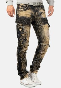 Jeans strappati con una combinazione di colori nero e tan, caratterizzati da borchie metalliche, zip e una vestibilità affusolata. Abbinati a sneakers bianche.