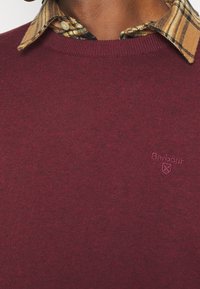 Pull en tricot bordeaux avec un col rond, arborant un logo brodé. Le col présente un motif à carreaux en beige et couleurs foncées.