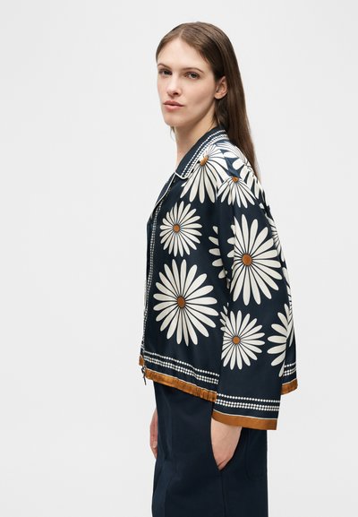 Γυναίκα με μακριά καστανά μαλλιά, που φοράει σκούρο μπλε μπουφάν με μεγάλα λευκά σχέδια μαργαρίτας και καφέ περιθώριο, στέκεται σε προφίλ με το χέρι στην τσέπη.
