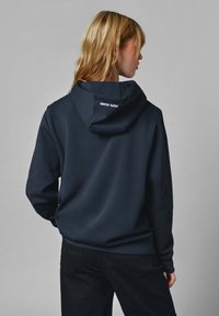 Sudadera de algodón azul marino con un ajuste relajado, capucha con cordón y detalle de marca en la parte delantera en blanco. Textura suave, mangas largas, diseño casual.