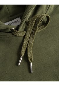Felpa verde oliva con tessuto testurizzato, caratterizzata da un cordoncino con estremità metalliche. Etichetta interna con la scritta "LES DEUX". Prospettiva ravvicinata.