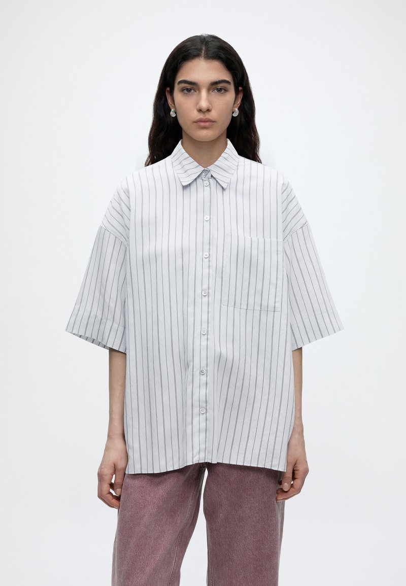 Jeune femme aux longs cheveux foncés porte une chemise blanche oversize à fines rayures verticales et un pantalon mauve sur fond uni.