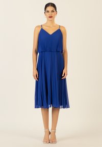 Faltenblaues Kleid mit dünnen Trägern, V-Ausschnitt und einem fließenden Rock. Gestylt mit silbernen Riemchenabsätzen. Glattes, leichtes Material.