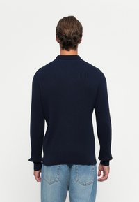 Man met kort bruin haar, gekleed in een navy geribbelde trui en lichtblauwe jeans, staande voor een effen witte achtergrond.