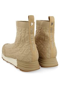 Botines beige de tobillo con tela texturizada que muestra un patrón repetido, con tiradores y una suela media blanca con acentos acanalados.