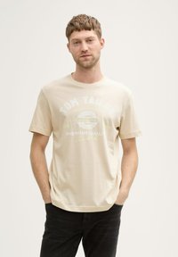 Bežs īsām piedurknēm T-krekls ar teksturētu apdari. Priekšpusē ir grafiskais teksts "TOM TAILOR" un "PREMIUM QUALITY" gaišās krāsās. Saskaņots ar tumšām džinsām.
