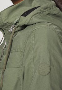 Olivengrøn jakke med tekstureret stof, lynlås, snøre i hætten og et rundt logo-mærke på ærmet. Har sorte netdetaljer.