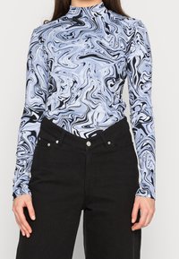 Haut à manches longues dans un motif tourbillon bleu et noir, présentant un col montant et une texture côtelée, associé à un jean noir avec des éléments de quincaillerie visibles.