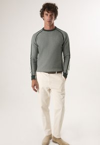 Grijze gebreide trui met groene accenten en witte gestreepte details, gecombineerd met crèmekleurige broek en tan boots.