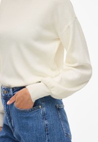 Cream knit sweater with ribbed cuffs, worn with high-waisted blue denim jeans featuring a classic five-pocket design and metal button closure.