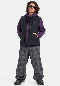 Zwarte mouwloze vest over een paarse hoodie, gecombineerd met grijze geruite wijde broekspijpen en zwarte sneakers, met een ontspannen pasvorm en zichtbare zakken.