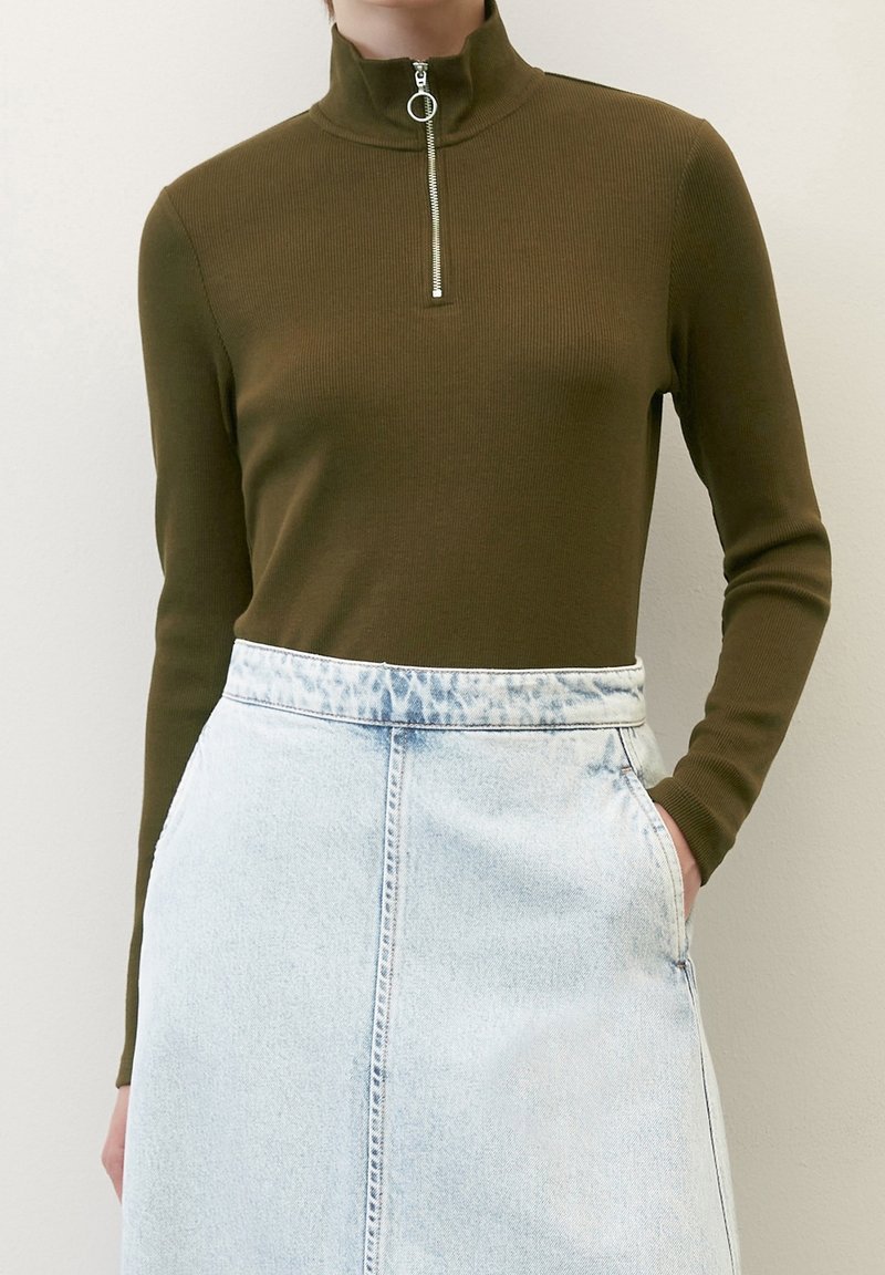 Personne portant un haut à manches longues côtelé vert olive avec un col zippé argenté et une jupe en denim taille haute bleu clair, la main dans la poche.