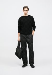 Juodas megztas megztinis su ląstelių raštu, derinamas su laisvomis juodomis kelnėmis, ant kurių subtiliai matomi lapų raštai. Laiko juodą siuvinėtą rankinę.