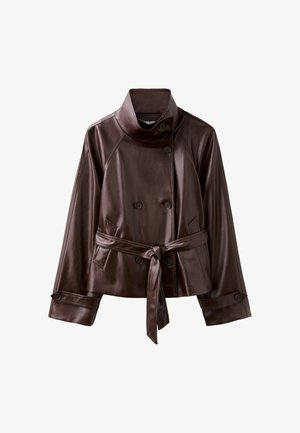 Femme portant une veste en cuir marron foncé ceinturée, un short en jean bleu clair longueur genoux, et des sandales blanches à talons, debout sur un fond beige.
