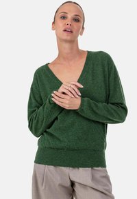 Grüner Strickpullover mit V-Ausschnitt, weiter Passform und geripptem Saum. Verfügt über lange Ärmel und eine weiche Textur, getragen über hellgrauer Hose.