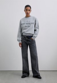 Sudadera gris con el texto "SPORT HERITAGE", combinada con vaqueros negros de tiro alto y pierna ancha. Tejido suave, ajuste relajado y diseño minimalista.