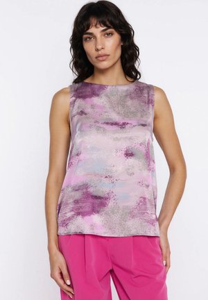 Kobieta o falowanych ciemnych włosach, ubrana w top bez rękawów z różowo-fioletowo-szarym abstrakcyjnym wzorem, w połączeniu z jaskraworóżowymi spodniami, stojąca na białym tle.