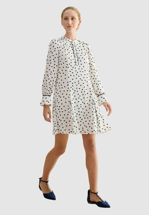 Mujer caminando con un vestido blanco de manga larga con lunares negros y zapatos planos puntiagudos azul marino con correas en el tobillo y adornos decorativos.