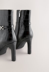 Pair of black faux crocodile leather ankle boots with high block heels and side zippers on a light background.