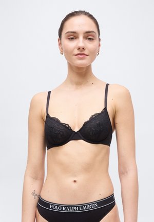 Kvinde iført sort blonde-bh og sort Polo Ralph Lauren undertøj, stående mod en ensartet lys baggrund, med en tatovering på venstre arm.