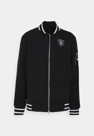 Veste bomber noire zippée avec col, poignets et ourlet à rayures blanches, affichant le logo des Raiders sur la poitrine et un swoosh Nike sur la poche de la manche.
