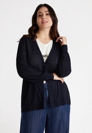 Cardigan marine avec un devant à boutons, des manches plissées texturées et des poignets côtelés. Porté sur un haut blanc avec un design d�écoratif.