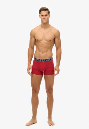 Shorts de boxers rojos de algodón con cintura elástica gris etiquetada como "SUPERDRY," usados por un modelo masculino de pie, descalzo, sobre un fondo simple.