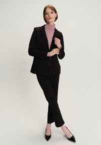 Zwart op maat gemaakt colbert over een lichtroze coltrui, gecombineerd met zwarte slim-fit broek en puntige zwarte hakken, tegen een lichte achtergrond.