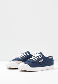 Marineblaue Canvas-Sneaker mit weißen Gummisohlen, weißen Schnürsenkeln, einer strukturierten Zehenkappe und einem hellen Akzent an der Seite.