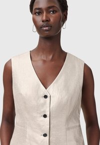 Ljust beige ärmlös klänning med djup V-krage, med en slät textur, fyra svarta knappar och sidofickor.