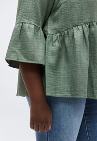 Κοντινό πλάνο σκουρόχρωμου χεριού που φοράει πράσινο μπλουζάκι με υφή και μανίκια καμπάνα, μαζί με μπλε τζιν τζιν παντελόνι, σε απλό ανοιχτό φόντο.