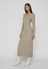 Ribbed, long-sleeve, beige knit dress with a rounded neckline, reaching the ankles, paired with shiny, cream-colored knee-high boots.