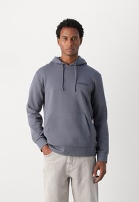 Siva kapuca s puloverjem s sprednjim žepom in vrvico. Ima majhen logotip na prsih in rebraste manšete. Obuto z svetlo obarvanimi kavbojkami.