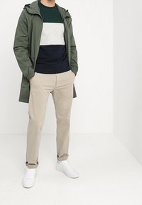 Hombre vestido con chaqueta verde con capucha, suéter a rayas, pantalones beiges y zapatillas blancas, de pie con las manos en los bolsillos.