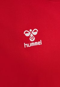 Rød T-shirt lavet af blødt stof med rund halsudskæring, med en hvid logo, der ligner en bi, og mærkenavnet "hummel" under.