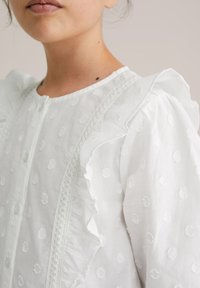 Blouse blanche à pois texturés, manches à volants et bordure en dentelle. Fermeture à boutons à l'avant. Tissu doux et léger.