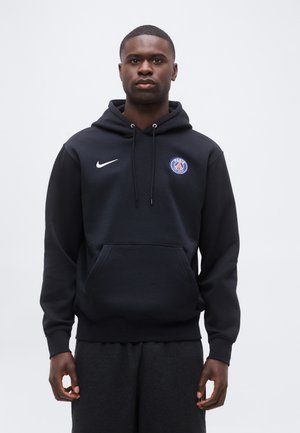 Homme portant un sweat à capuche noir Nike avec le logo du Paris Saint-Germain, debout devant un fond uni gris clair, regardant devant.