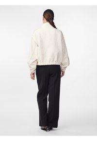 Kremowa, pikowana kurtka bomberka z wysokim kołnierzem i elastycznym pasem, w zestawie z czarnymi spodniami o szerokich nogawkach. Gładka tekstura i luźny krój.