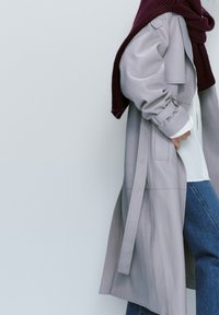 Lunga trench grigia con ampi risvolti e bottoni, sovrapposta a una camicia bianca. Accentuata da una sciarpa a coste bordeaux. Pantaloni in denim casual.