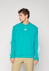 Young man in teal Adidas sweatshirt with white stripes on sleeves, beige pants, looking to his left against plain gray background.