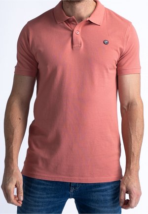 Homme portant un polo corail à manches courtes avec deux boutons et un petit logo rond sur la poitrine, assorti à un jean bleu.