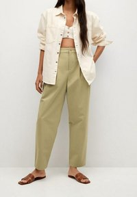 Chemise boutonnée beige clair portée sur un crop top blanc, assortie à un pantalon taille haute vert olive et des sandales à glissière marron.