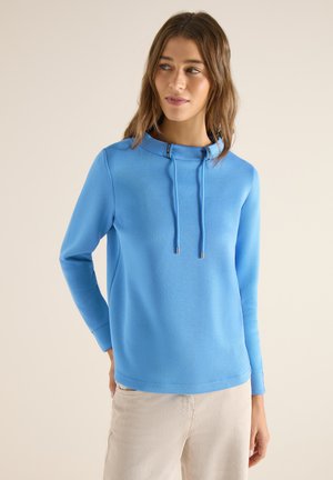 Młoda kobieta w jasnoniebieskiej bluzce z długim rękawem i sznurkami oraz beżowych spodniach, patrząca lekko na bok na jednolitym tle.