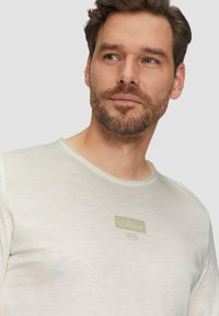 Jasno beżowa koszulka z okrągłym dekoltem, wykonana z miękkiego materiału, z prostokątną naszywką z logo w zieleni i bieli na lewej stronie klatki piersiowej.