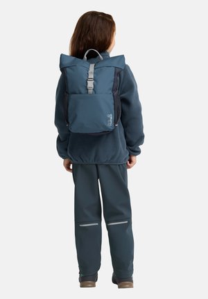 Zaino in tessuto blu con parte superiore pieghevole, spallacci grigi e tasca frontale in rete, indossato sopra una giacca in pile e pantaloni grigi.